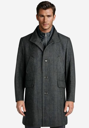 Mann trägt einen dunkelgrauen Mantel mit Fischgrätmuster und einer schwarzen Reißverschluss-Innenschicht, steht vor einem schlichten weißen Hintergrund.