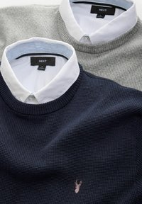 To sweatere med krave: en marineblå med et tekstureret mønster og et lille broderet pink logo, den anden lysegrå med en glat overflade.