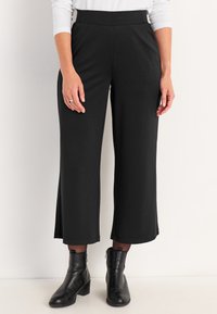 Zwarte, wijde broek met een gladde textuur, hoge tailleband en zijsplitten; gecombineerd met zwarte enkellaarzen.