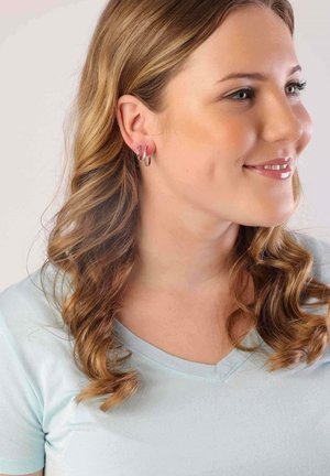 Des boucles d'oreilles en argent en forme de créole, avec une finition lisse et polie, portées par une personne ayant des cheveux ondulés de longueur épaule, vêtue d'une chemise bleu clair.