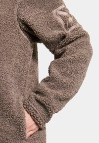 Brun fleece sweater med en blød, tekstureret overflade; har et fremtrædende logo på ærmet og en sidelomme.