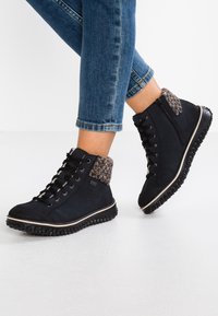 Bottes montantes noires avec un revers en tissu texturé, un laçage à l'avant et une semelle en caoutchouc avec un motif de crampons. Zippées sur le côté pour un enfilage facile.