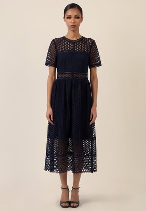 Frau in einem marineblauen, kurzärmligen Spitzenkleid in knielanger Länge mit transparentem Muster, stehend vor einem schlichten beigefarbenen Hintergrund.