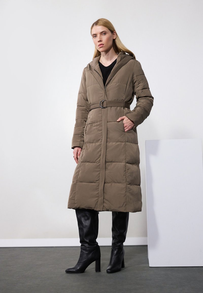 Manteau long rembourré en vert olive avec une taille ceinturée, une capuche et un motif de coutures en diagonale. Porté avec des bottes noires en cuir à talons hauts.