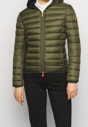 Person iført en tætsiddende olivengrøn quiltet dynejakke med hætte, frontlynlås med orange trækflige, kombineret med sorte bukser.