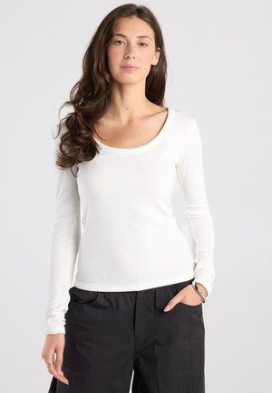Jonge vrouw met lang bruin haar draagt een aansluitend wit shirt met lange mouwen en zwarte broek, staand met één hand in de zak tegen een effen achtergrond.