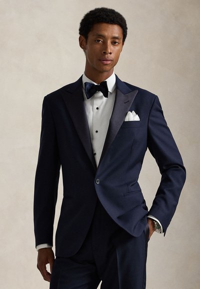 Man wearing a navy blue tuxedo with satin lapels, white dress shirt, black bow tie, and white pocket square, standing with one hand in pocket.