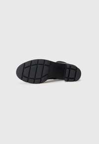 Semelle de chaussure noire avec une surface en caoutchouc texturée, présentant un motif en grille, une forme arrondie et une finition lisse. Logo de marque notable présent.