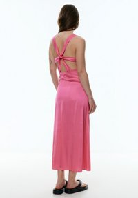 Rosa Satin-Maxikleid mit einem verdrehten Rückendesign, breiten Trägern und einer glatten Textur. Getragen mit schwarzen Sandalen.