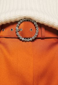 Oranje stoffen broek met een ronde zilveren gesp versierd met heldere rhinestones, gecombineerd met een crèmekleurige geribbelde trui bij de taille.