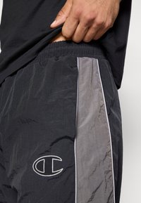 Persoon die zwart-grijze sportbroek met elastische tailleband en een logo op de linkerdij optrekt.