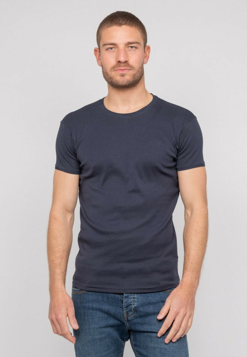 T-shirt ajusté bleu marine avec des manches courtes, un col rond et une texture lisse. Présente un petit logo sur le devant, associé à un jean bleu.