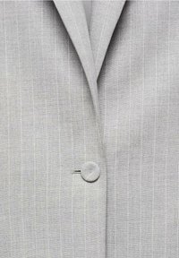 Vaaleanharmaa bleiseri, jossa on teksturoitu kangas pystysuorilla raidallisilla yksityiskohdilla ja pyöreällä nappisuljennalla, korostaen räätälöityä muotoilua.