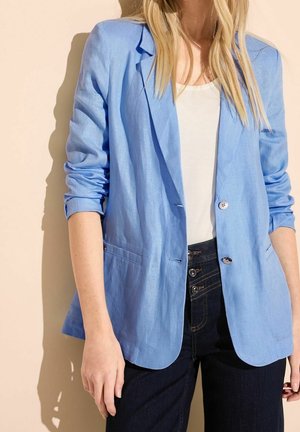 Vrouw met een lichtblauw colbert met opgerolde mouwen, een wit topje en een high-waisted donkere spijkerbroek met meerdere knopen.