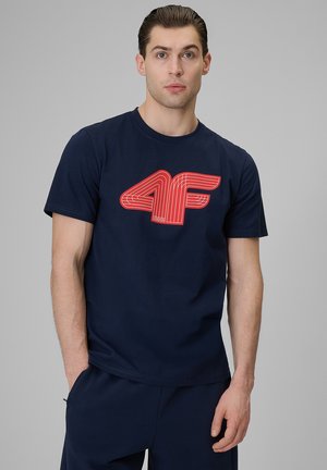 Młody mężczyzna z zaczesanymi do tyłu włosami, ubrany w granatowy t-shirt z dużym czerwonym logo „4F”, stoi na jednolitym szarym tle.