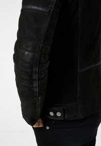 Schwarze Lederjacke mit strukturierter Oberfläche, gesteppte Ellbogenpatches und silbernen Druckknopfdetails an der Taille und den Ärmeln. Mit Reißverschluss.