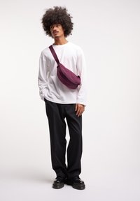 Borsa a tracolla bordeaux realizzata in tessuto strutturato, chiusura frontale con zip, indossata sulla spalla sinistra sopra una camicia bianca a maniche lunghe e pantaloni neri.
