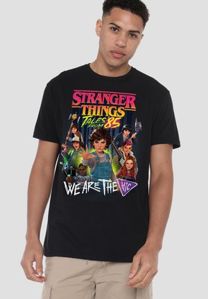 Junger Mann trägt ein schwarzes T-Shirt mit den Figuren aus "Stranger Things Tales from '85" und buntem Schriftzug "We Are The Vic".