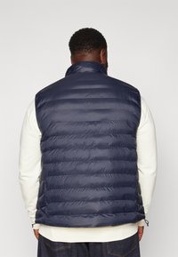 Man sett från ryggen iklädd en marinblå vadderad väst över en vit långärmad skjorta och mörka jeans mot en enfärgad bakgrund.