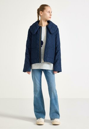 Námořnicky modrá prošívaná bunda s velkým límcem, zlatým zipem a předními kapsami, vrstvená přes šedý svetr a doplněná rozšířenými džínami a teniskami.