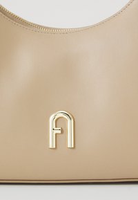 Bēšs ādas rokassoma ar gludu tekstūru, noapaļotu formu un zelta toņa metāla logotipa akcentu priekšpusē. Ar rāvējslēdzēju aizverama augšdaļa.