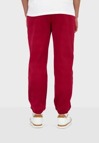Pantalones de chándal rojos hechos de una mezcla de algodón, con puños elásticos y un corte relajado. Llevados con zapatillas blancas, que muestran detalles en marrón.