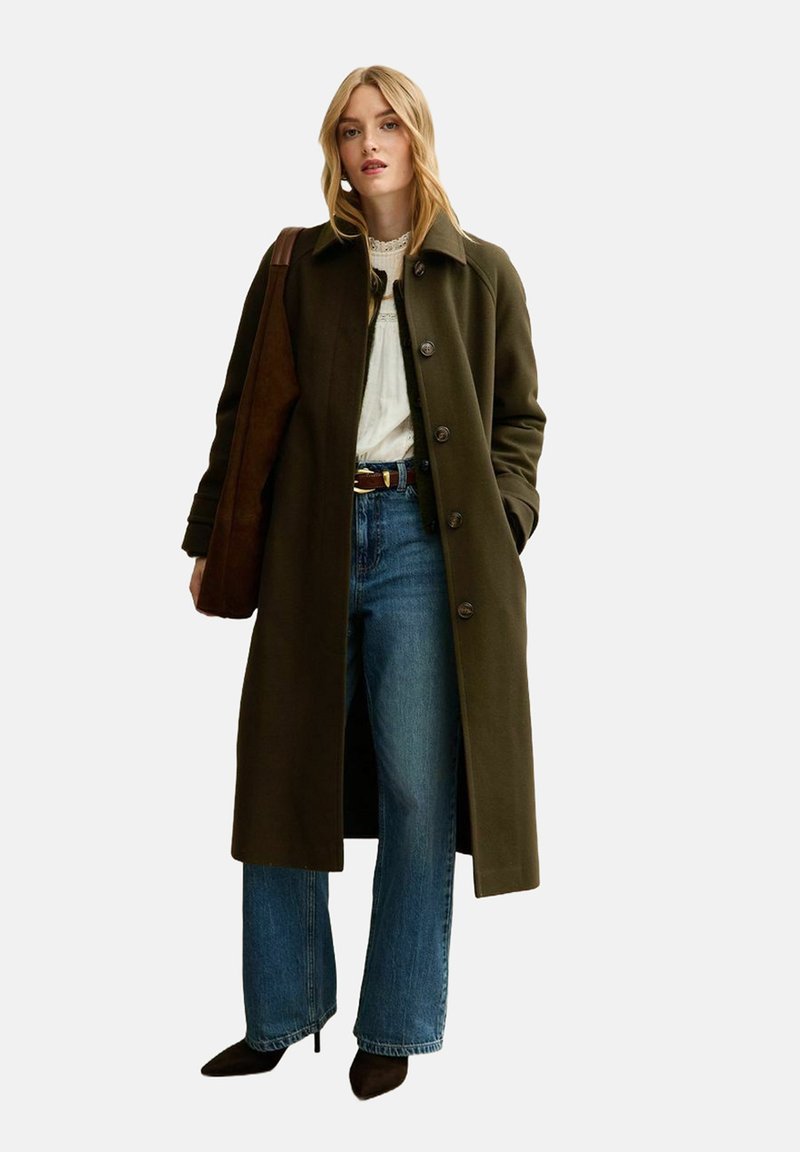 Abrigo largo de color verde oliva con grandes botones, usado sobre una blusa clara, combinado con jeans de tiro alto de color azul y botas de tobillo marrones.