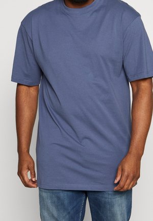 Man draagt een effen blauw T-shirt met korte mouwen en een blauwe spijkerbroek, staand tegen een neutrale lichte achtergrond.