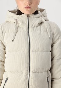 Veste matelassée beige en texture côtelée, avec capuche, fermeture éclair à l'avant et poches latérales. Les coutures incurvées ajoutent de la dimension au design.