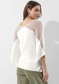 Witte top met doorzichtig, lichtgekleurde mesh mouwen, ronde halslijn en uitlopende design. Gecombineerd met olijfgroene broeken met brede pijpen.