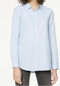 Chemise à manches longues bleu clair, confectionnée dans un tissu doux, avec un col et un ourlet arrondi. Elle se ferme par des boutons à l'avant et possède des poignets.