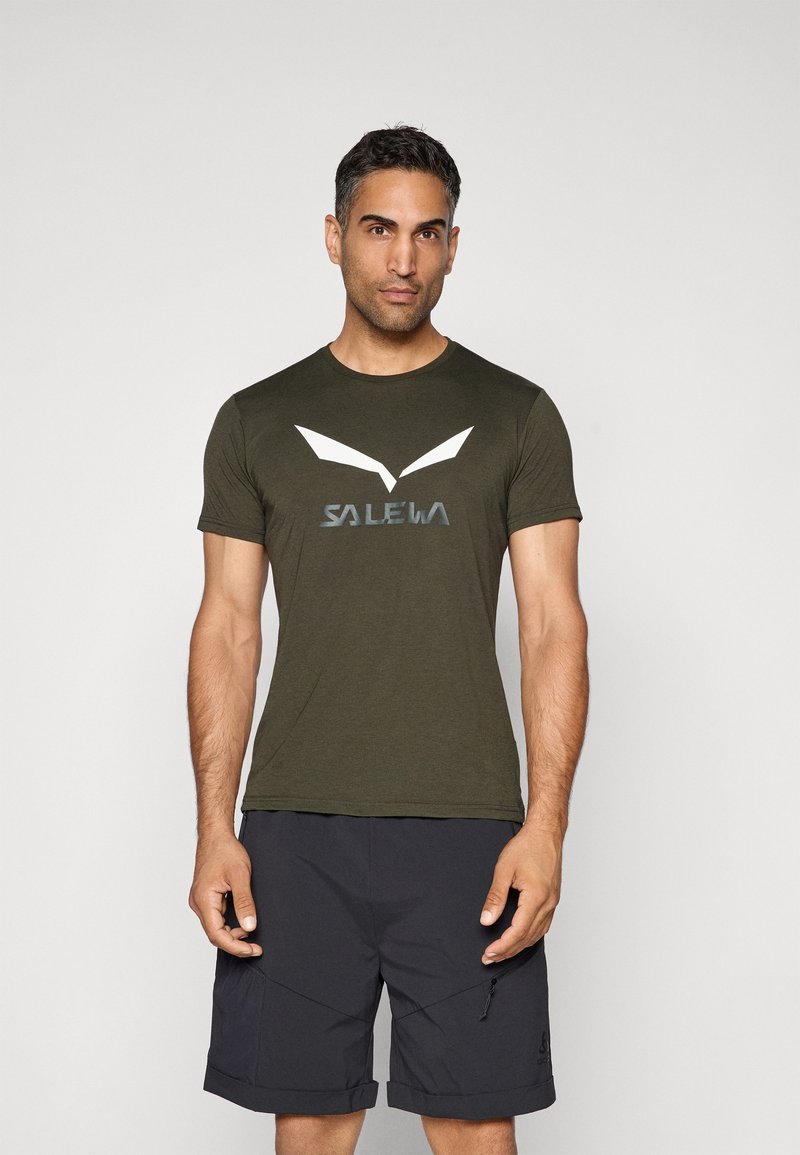 Olivengrønn T-skjorte med korte ermer, hvitt og grått logo, rund hals og glatt tekstur, sammen med mørke shorts.