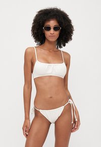 Hvit bikiniset med teksturert crop top med tynne stropper og lavt snitt på bunnen med sidesnøring. Minimalistisk design og nøytral farge.