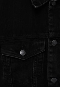 Veste en denim noire en gros plan montrant une poche à rabat avec un bouton en métal et un détail partiel du col.