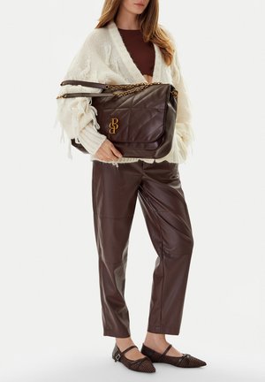 Mujer con cárdigan de punto color crema y pantalones de cuero marrón sosteniendo un gran bolso de cuero marrón oscuro con asas de cadena dorada.