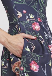 Kukallinen mekko, jossa on tumma tausta ja kirkkaat värit. Mekossa on kengurukuoppa ja pehmeä, joustava kangas.