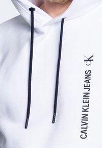 Sudadera blanca con capucha, con cordones negros y "CALVIN KLEIN JEANS" impreso en vertical en negro en el lado derecho.