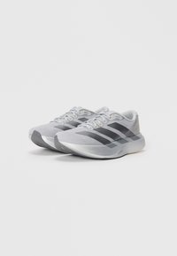 Chaussures de sport grises avec dessus en mesh, trois rayures noires, bout arrondi et semelle texturée. Conception légère avec des accents réfléchissants.