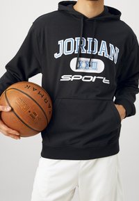 Felpa nera con tasca frontale, con testo bianco e blu "JORDAN XXIII" e "SPORT." Tiene un pallone da basket, indossata con pantaloni corti bianchi.