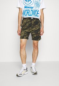 Personne portant un t-shirt graphique blanc, un short cargo camouflé, des chaussettes blanches et des chaussures de course blanches avec des détails jaunes et noirs.