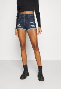 Tmavě modré džínové kraťasy s efektem opotřebení, ohrnutými lemy a standardním pasem. Kombinovány s černými šněrovacími bojovými botami.