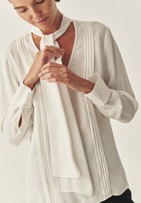 Biała bluzka z pionowymi fałdami i wiązaniem w formie chusty. Wykonana z miękkiego materiału, z długimi rękawami i zapięciem na guziki przy nadgarstkach.