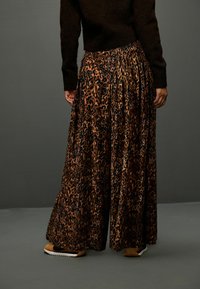 Kalhoty se širokými nohavicemi a plisováním v leopardím vzoru, vyrobené z hnědé a černé sametové látky, doplněné teniskami a elastickým pasem.