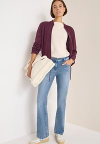 Chaqueta de cremallera color burdeos, suéter de punto beige, jeans acampanados de un azul claro, manta de felpa color crema, y zapatillas beige con detalles azules.