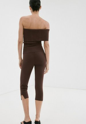 Femme aux cheveux foncés attachés en chignon, portant un crop top marron à épaules dénudées et un pantalon court assorti et ajusté avec de petites fentes aux jambes, debout dans une pièce blanche.