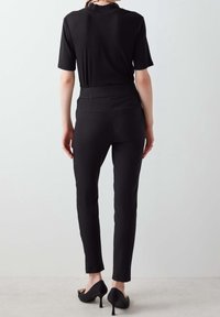 Osoba stojí zády k divákovi, na sobě má černý krátký rukáv a přiléhavé černé kalhoty, které doplňují vysoké podpatky.