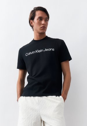 Νέος άνδρας με μαύρο t-shirt Calvin Klein Jeans και λευκό παντελόνι με υφή, στέκεται με τα χέρια στις τσέπες, κοιτάζοντας στο πλάι.