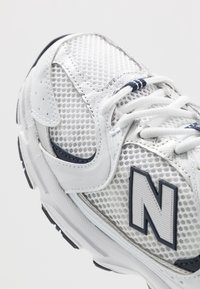 Vit idrottssko med meshöverdel, svart och grå "N"-logotyp, vita snören och vadderad sula, visad i närbild från en vinklad vy.