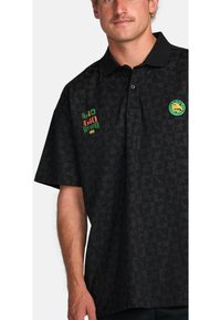 Black polo shirt with star pattern, featuring a green and red logo on the chest and a circular badge on the sleeve. Short sleeves, relaxed fit.