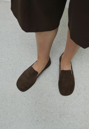 Legs wearing dark brown suede slip-on shoes and matching dark brown skirt standing on light textured floor.